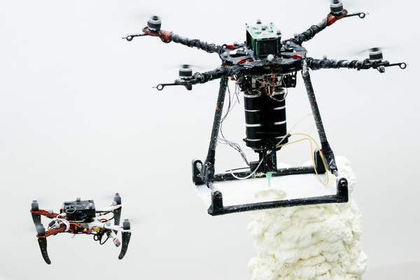 Two drone-like robots. A smaller one is on the left. A larger one is on the right. The larger one is making a 3D printout of something that looks like white foam. 